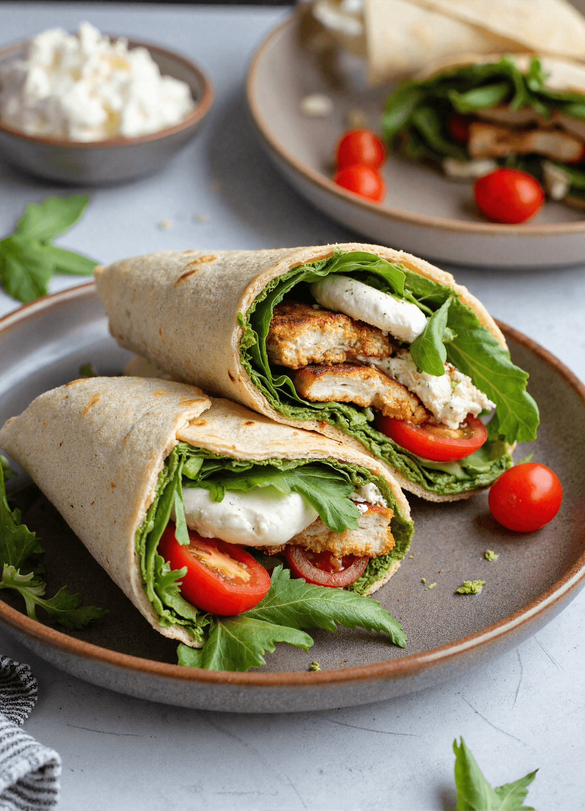 Sliced chicken pesto wraps with mozzarella, cherry tomatoes, and leafy greens on a brown ceramic plate, garnished with fresh herbs.