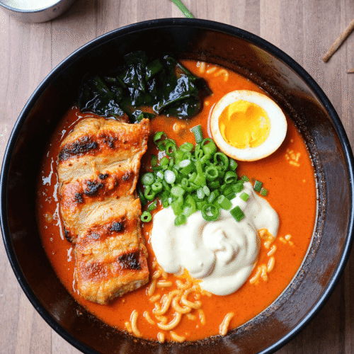 Bowl of fiery chicken ramen with grilled chicken, soft-boiled egg, creamy garlic sauce, scallions, and seaweed in spicy broth.