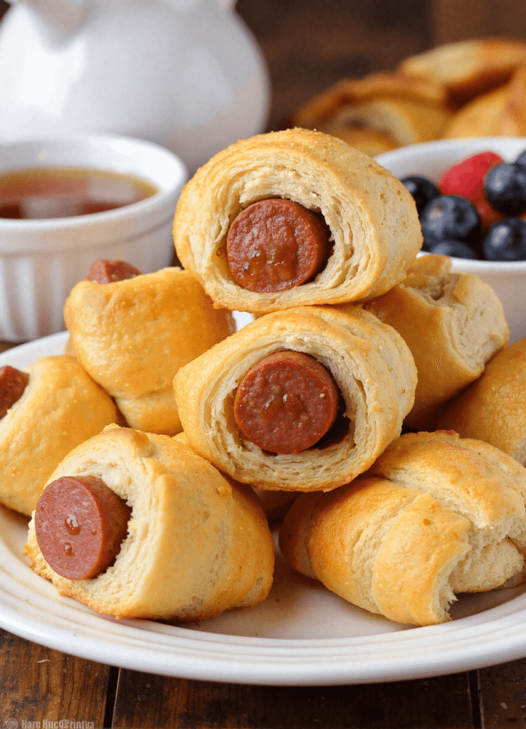 Plate of freshly baked breakfast pigs in a blanket, golden brown crescent rolls wrapped around savory sausage, served with fruit and syrup on the side.