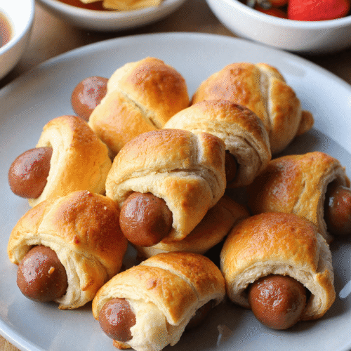 Freshly baked breakfast pigs in a blanket served on a white plate with syrup and fresh berries, showcasing a kid-friendly make-ahead morning treat.