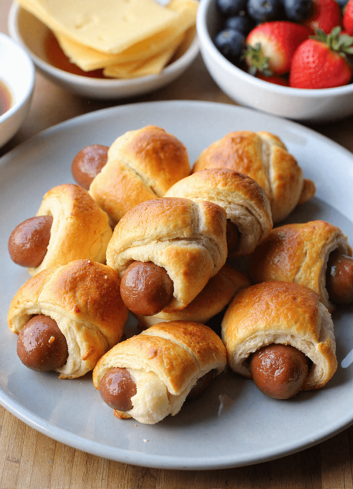 Freshly baked breakfast pigs in a blanket served on a white plate with syrup and fresh berries, showcasing a kid-friendly make-ahead morning treat.