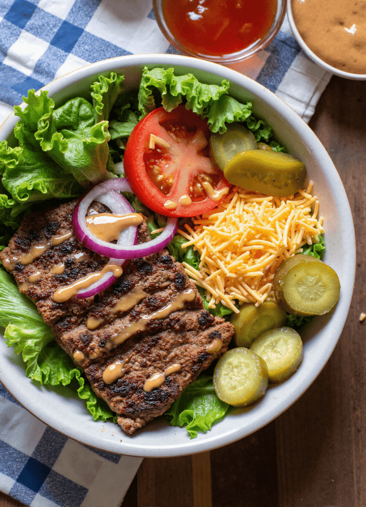 Burger bowl with grilled beef mini patties, cheddar cheese, pickles, tomato slices, onions, and low-carb burger sauce on fresh greens.