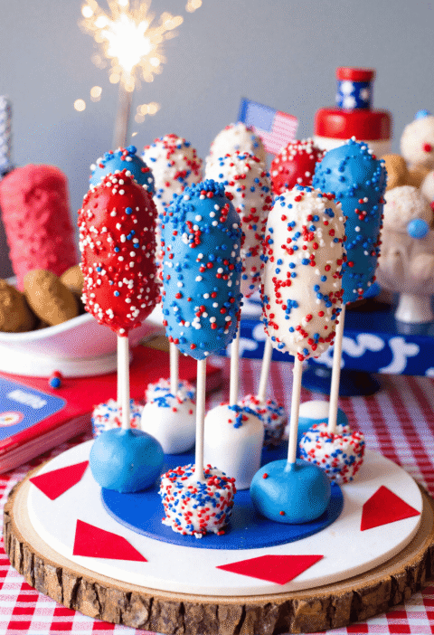 Patriotic Firecracker Rice Krispies Pops dipped in red, white, and blue candy melts, displayed upright with sprinkles on a circular stand with a sparkler in the background