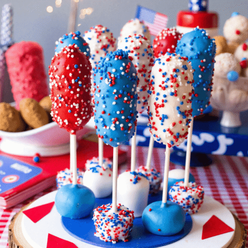 Patriotic Firecracker Rice Krispies Pops dipped in red, white, and blue candy melts, displayed upright with sprinkles on a circular stand with a sparkler in the background