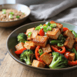 A vibrant bowl of garlic soy tofu stir-fry featuring crispy tofu cubes, broccoli florets, red bell peppers, and scallions served in a dark ceramic bowl.