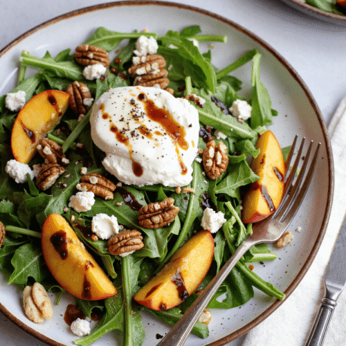 Grilled peach and burrata arugula salad with balsamic glaze, toasted pecans, and arugula on a rustic white plate