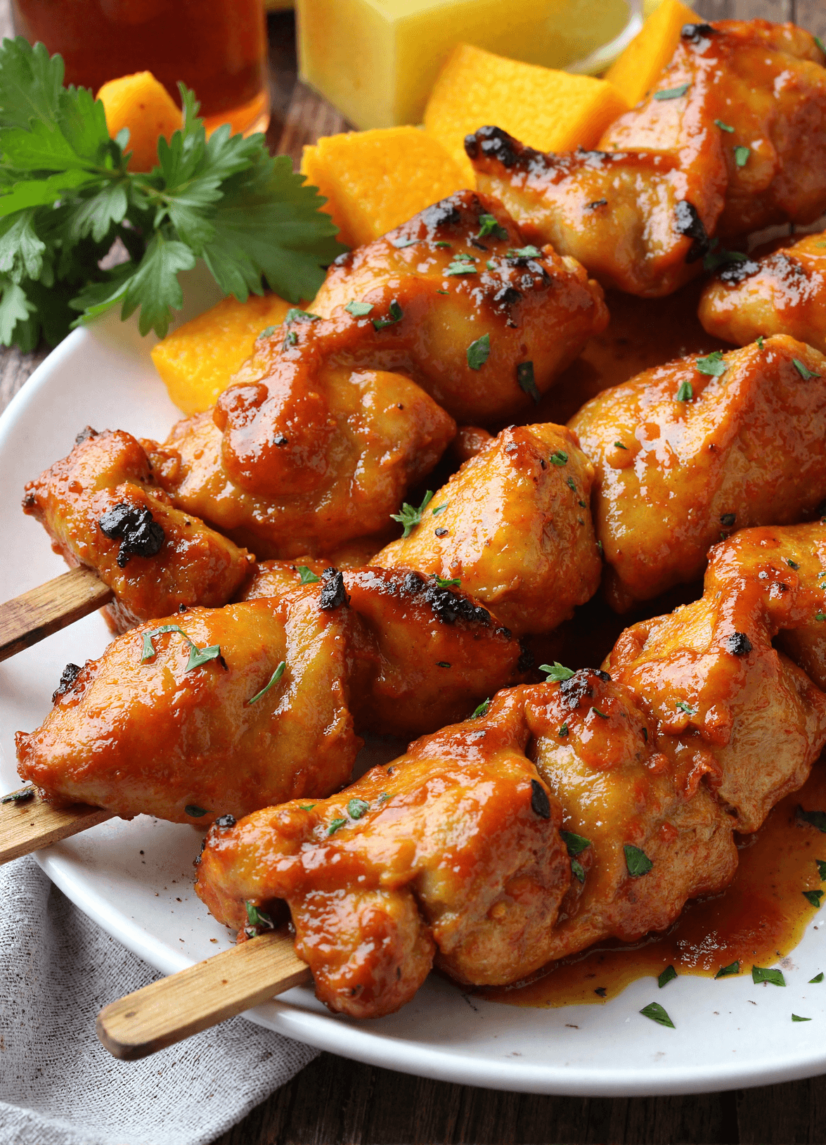 Close-up of grilled honey chipotle chicken skewers with golden caramelized glaze, served with fresh herbs and orange slices on a white plate.