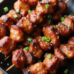 Charred pork skewers brushed with sticky honey garlic glaze and sprinkled with sesame seeds and scallions on a black round plate.