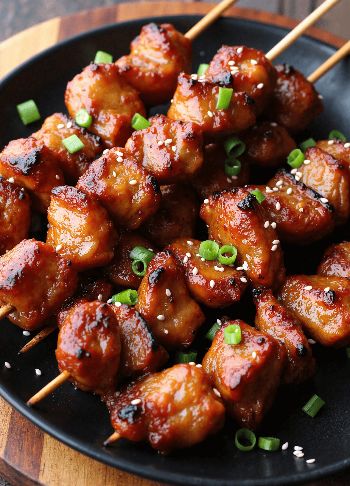 Charred pork skewers brushed with sticky honey garlic glaze and sprinkled with sesame seeds and scallions on a black round plate.