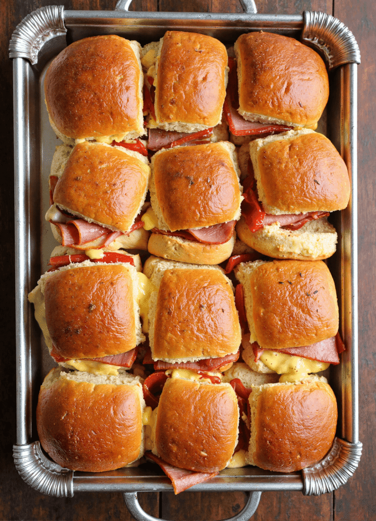 Baked hot Italian sub sliders layered with salami, ham, and provolone on Hawaiian rolls in a silver tray, fresh from the oven.