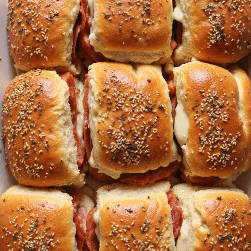 Baked Italian grinder sliders with melted cheese, deli meats, and sesame seed buns on a sheet pan.