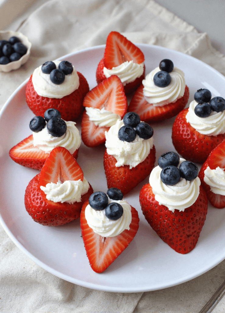 Fourth of July cheesecake stuffed strawberries arranged on a white plate and topped with blueberries and piped cream cheese filling