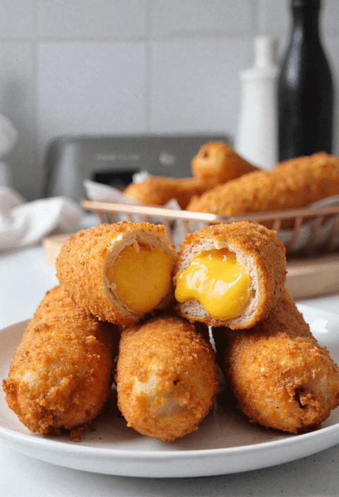 Korean corn dogs stacked on a plate with one cut open to reveal melted cheddar cheese, basket of more corn dogs blurred in the background.