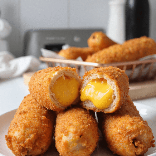 Korean corn dogs stacked on a plate with one cut open to reveal melted cheddar cheese, basket of more corn dogs blurred in the background.
