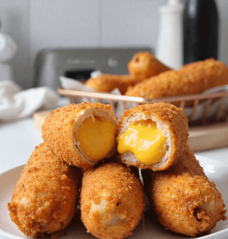 Korean corn dogs stacked on a plate with one cut open to reveal melted cheddar cheese, basket of more corn dogs blurred in the background.