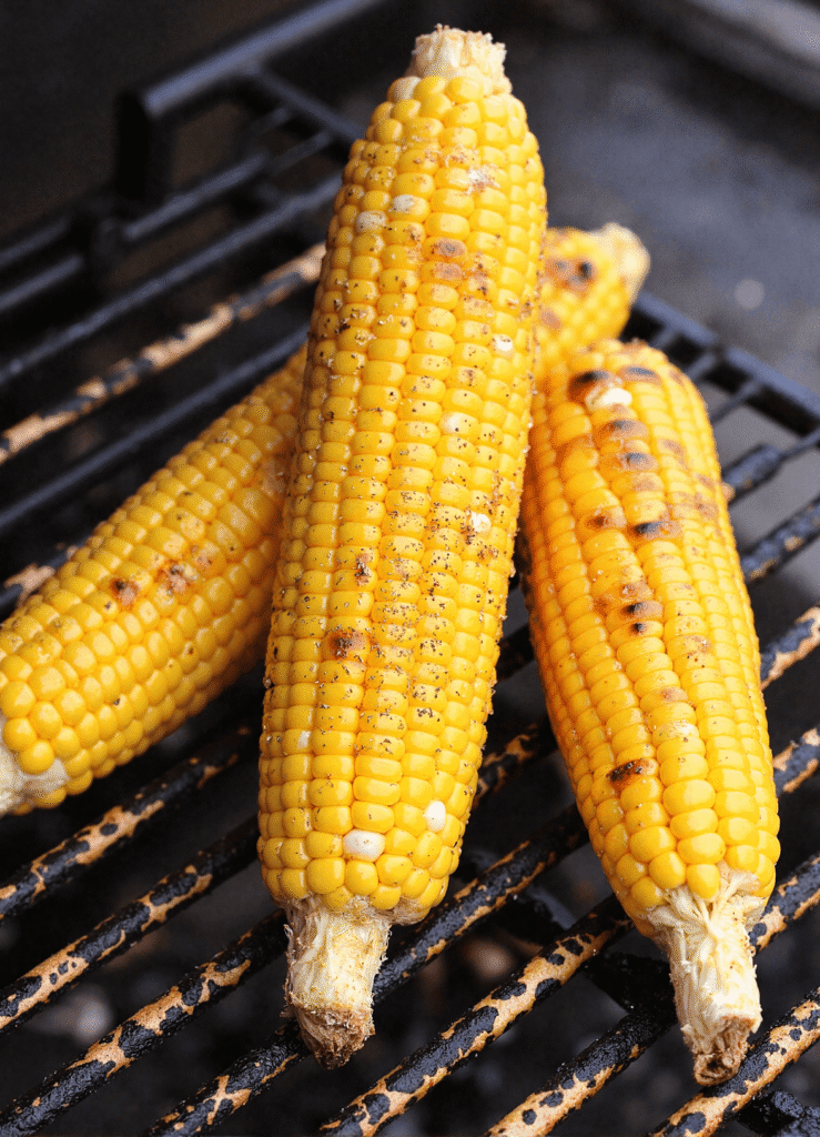 Three ears of lemon pepper corn grilling over open flames with blackened grill marks.