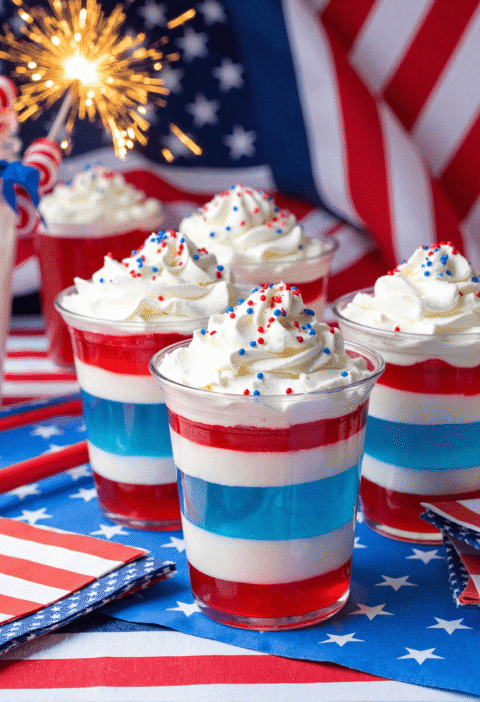 Red, white, and blue patriotic Jello layer cups topped with whipped cream and sprinkles in front of American flags and sparklers.