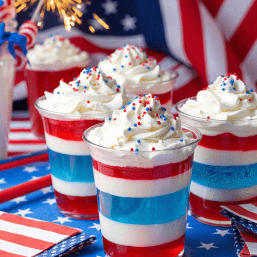 Red, white, and blue patriotic Jello layer cups topped with whipped cream and sprinkles in front of American flags and sparklers.