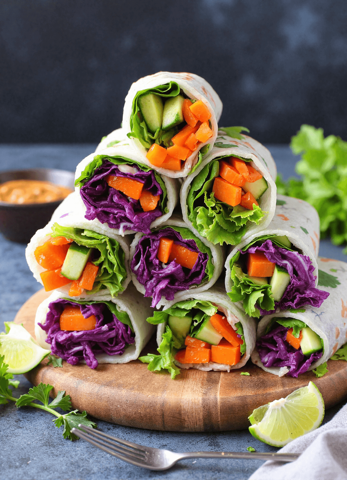 Stack of rainbow veggie rice paper rolls filled with purple cabbage, carrots, cucumber, and lettuce on a wooden board with lime wedges