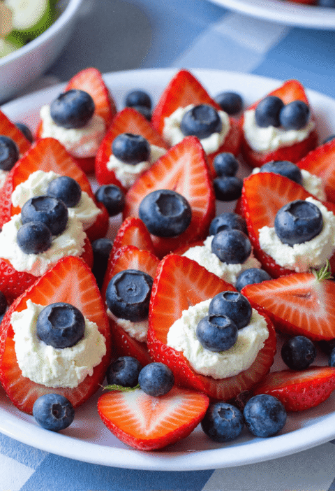 Plate of red, white & blue cheesecake stuffed strawberries topped with blueberries on a white dish, perfect for July 4th dessert tables