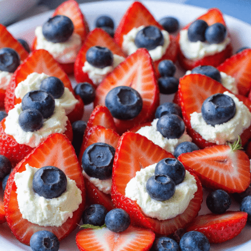 Plate of red, white & blue cheesecake stuffed strawberries topped with blueberries on a white dish, perfect for July 4th dessert tables