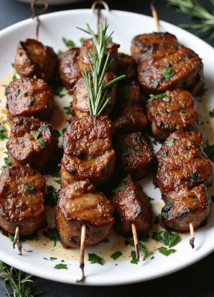 Juicy rosemary garlic steak kebabs grilled to perfection on skewers with fresh herbs on a white plate.