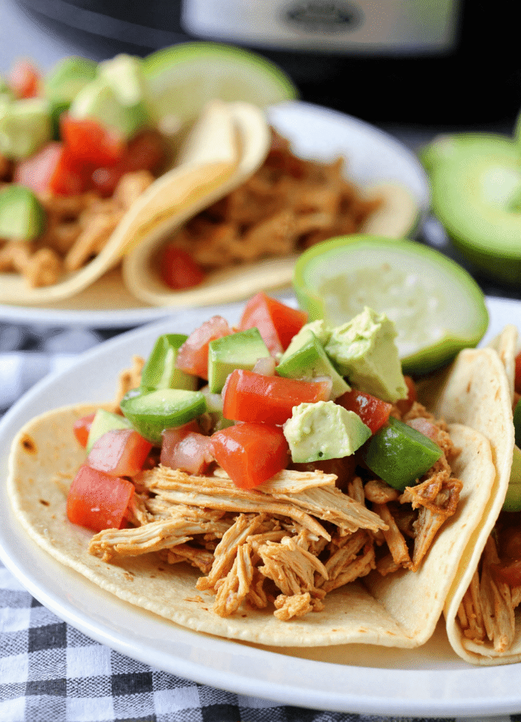 Close-up of slow cooker salsa chicken tacos topped with avocado, tomato, and lime wedges on a white plate