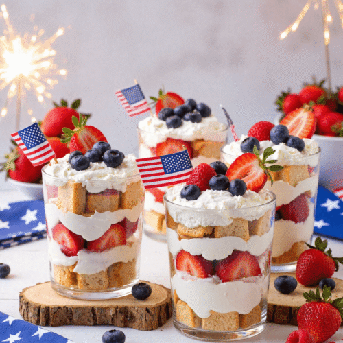 Individual Star-Spangled Berry Shortcake Trifles in glass cups, layered with whipped cream, strawberries, blueberries, and pound cake, topped with mini American flags and sparklers.