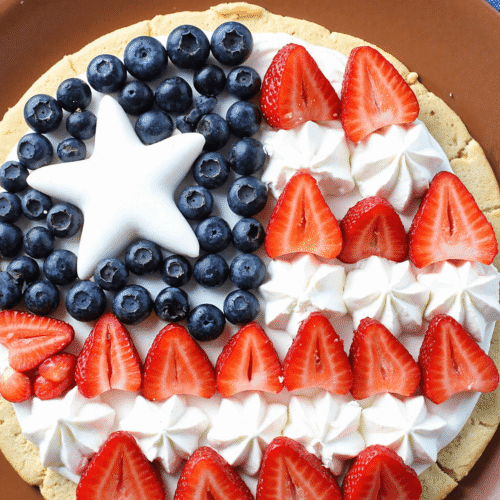 Patriotic stars and stripes fruit pizza topped with blueberries, strawberries, and piped whipped cream stars on a sugar cookie crust.