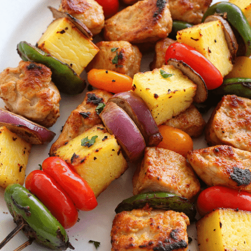 Grilled yummy honey chicken kabobs with pineapple, bell peppers, red onion, and jalapeño on metal skewers served on a white plate.