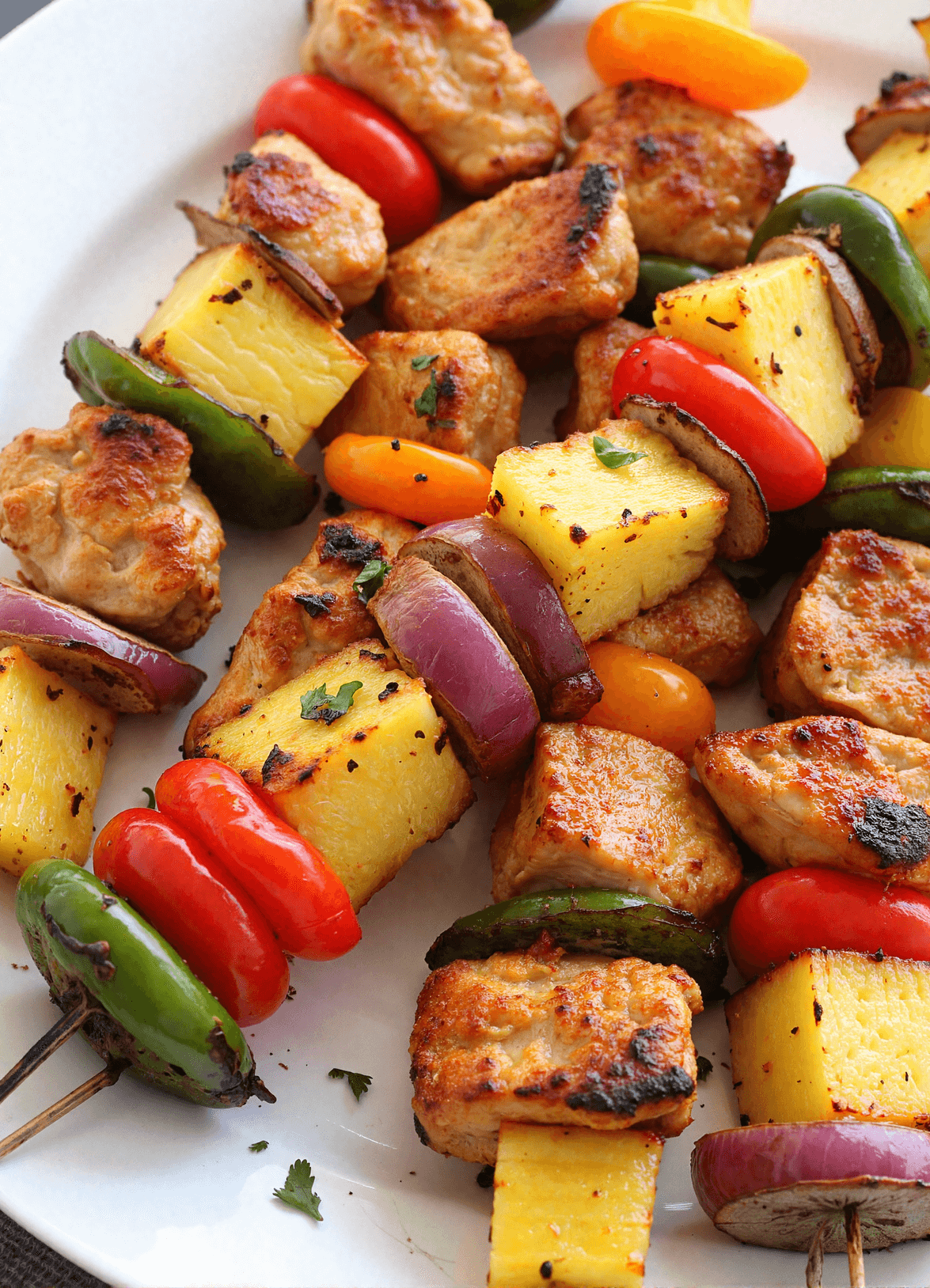 Grilled yummy honey chicken kabobs with pineapple, bell peppers, red onion, and jalapeño on metal skewers served on a white plate.