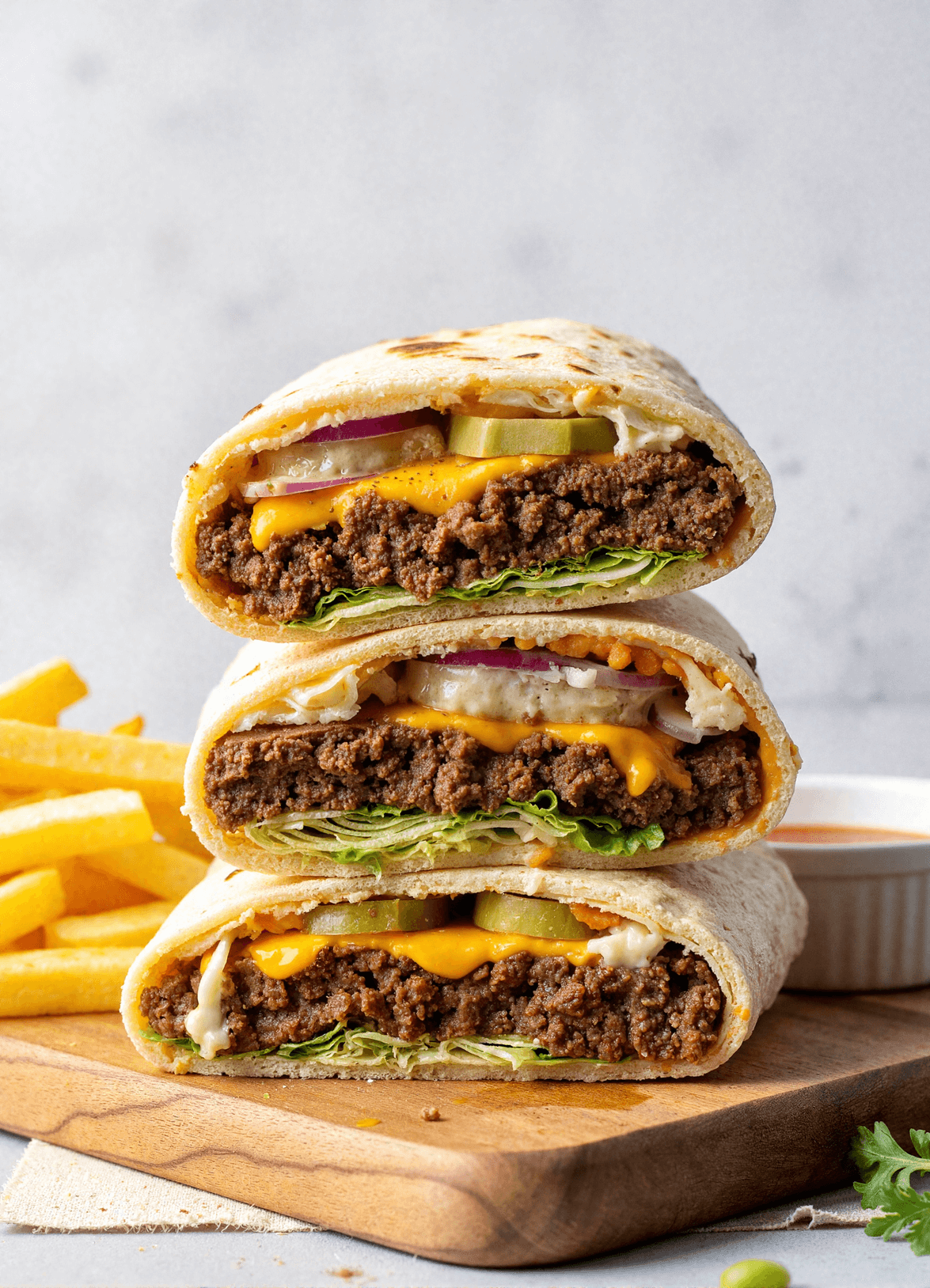 Cheesy Big Mac wraps stacked on a board, loaded with seasoned beef, cheddar cheese, pickles, onions, and lettuce, next to crispy fries.