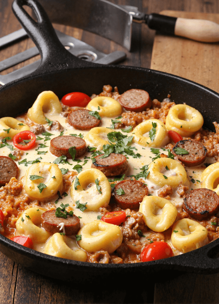 Cheesy Blackstone tortellini skillet with smoked sausage, creamy sauce, cherry tomatoes, and parsley in a cast iron pan.
