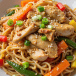 Chicken noodle stir fry with sliced chicken breast, bell peppers, sesame seeds, and green onions in a white bowl
