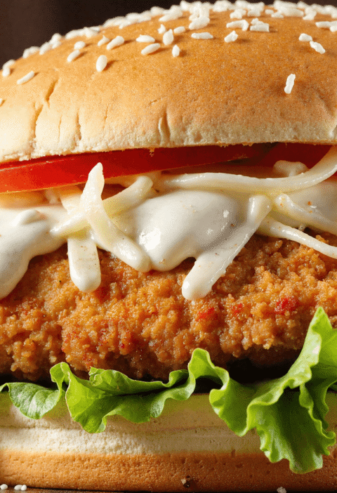 Close-up view of a crispy chicken Caesar sandwich with romaine lettuce, parmesan, Caesar dressing, tomato slices, and a sesame bun