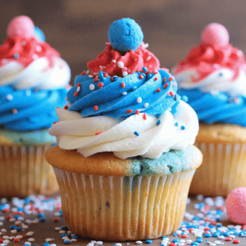 Red, white, and blue firecracker cupcake topped with patriotic frosting swirl and candy, decorated with sprinkles.
