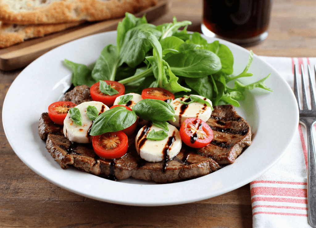 Grilled ribeye steak topped with sliced fresh mozzarella, ripe tomatoes, basil, and a drizzle of balsamic glaze.