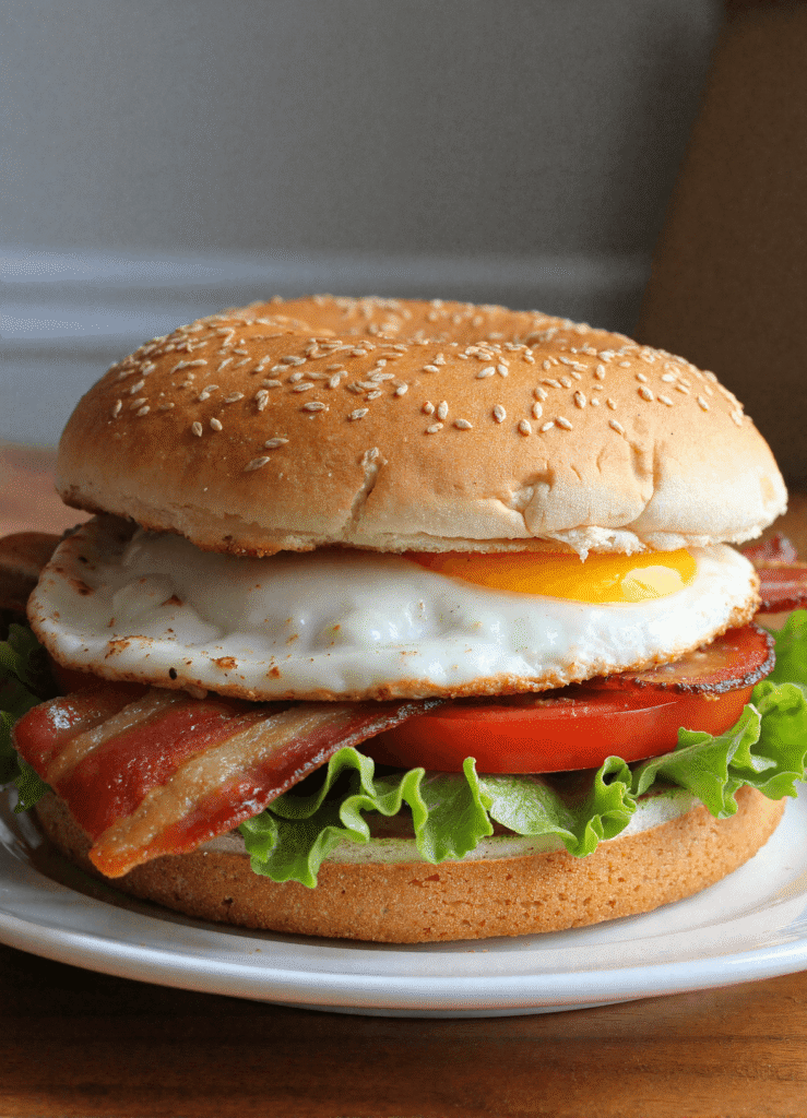 Stacked BLT breakfast sandwich on a sesame bagel with crispy bacon, sunny-side egg, lettuce, and tomato.