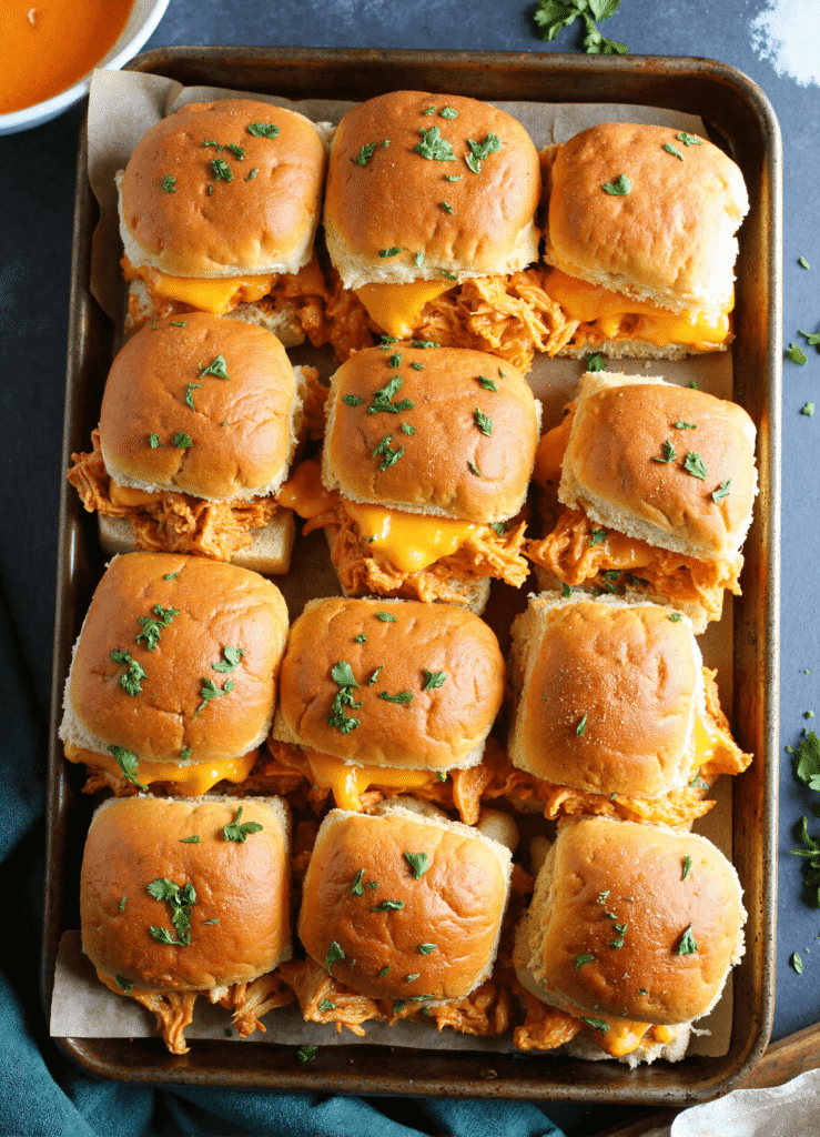 Overhead view of a baking tray filled with buffalo chicken sliders topped with cheddar and chopped parsley, perfect for party appetizers or easy weeknight meals.