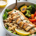 Grilled chicken and quinoa power bowl with roasted vegetables, fresh lemon wedge, and tahini drizzle in a white bowl.