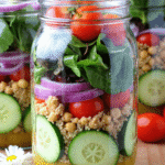 Layered cucumber jar salad with chickpeas, couscous, red onion, cherry tomatoes, and fresh greens in a glass mason jar.