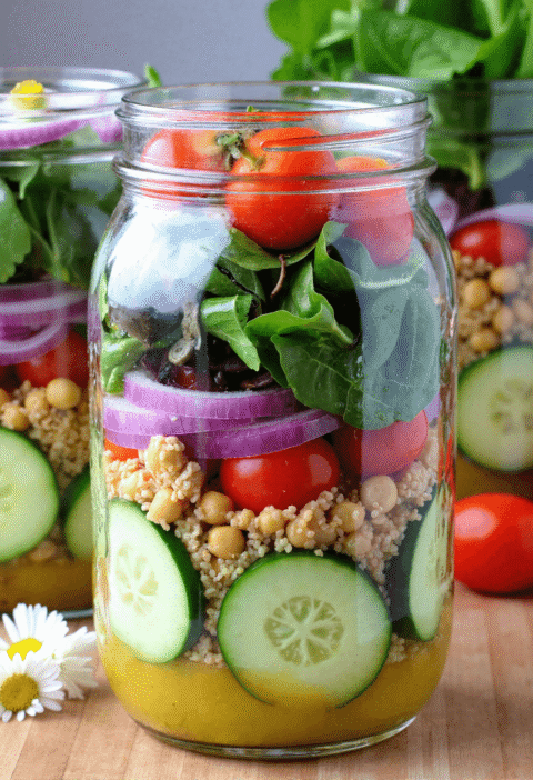 Layered cucumber jar salad with chickpeas, couscous, red onion, cherry tomatoes, and fresh greens in a glass mason jar.