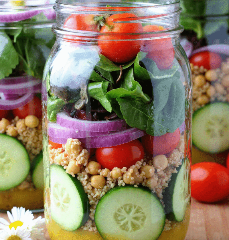 Layered cucumber jar salad with chickpeas, couscous, red onion, cherry tomatoes, and fresh greens in a glass mason jar.