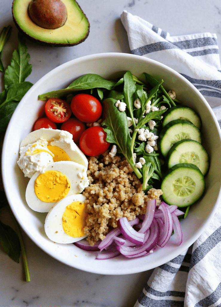 Fresh egg white power salad with spinach, quinoa, cherry tomatoes, sliced cucumber, avocado, and red onion in a white bowl