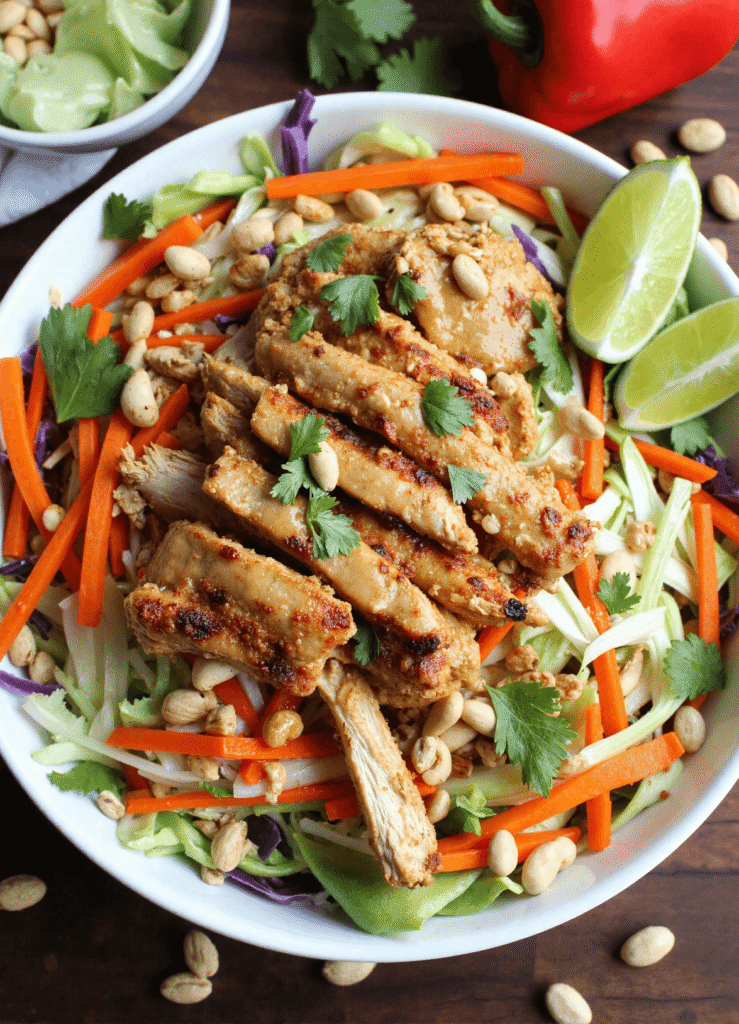 Sliced Thai peanut chicken served on a bed of shredded vegetables with cilantro, lime wedges, and crunchy peanuts in a white bowl.