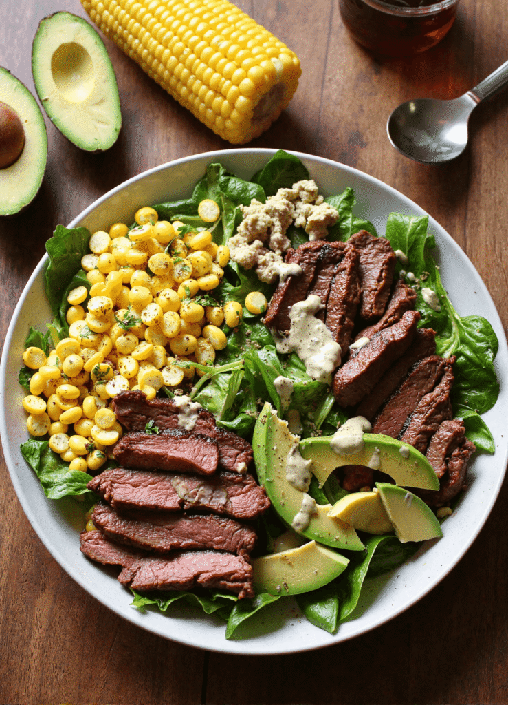 Grilled steak salad with avocado, roasted potatoes, corn, almonds, feta, and fresh vegetables on a white plate.