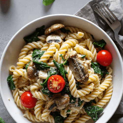 Top view of rotini pasta in a white bowl, tossed with sautéed mushrooms, spinach, and cherry tomatoes for a creamy plant-based vegan dinner.