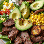 Grilled steak slices served on a bed of leafy greens with sweet corn, creamy avocado, and drizzle of garlic dressing.