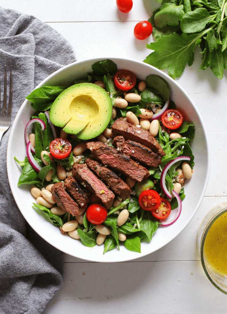 steak salad with white beans, avocado, tomatoes, and greens in a bowl