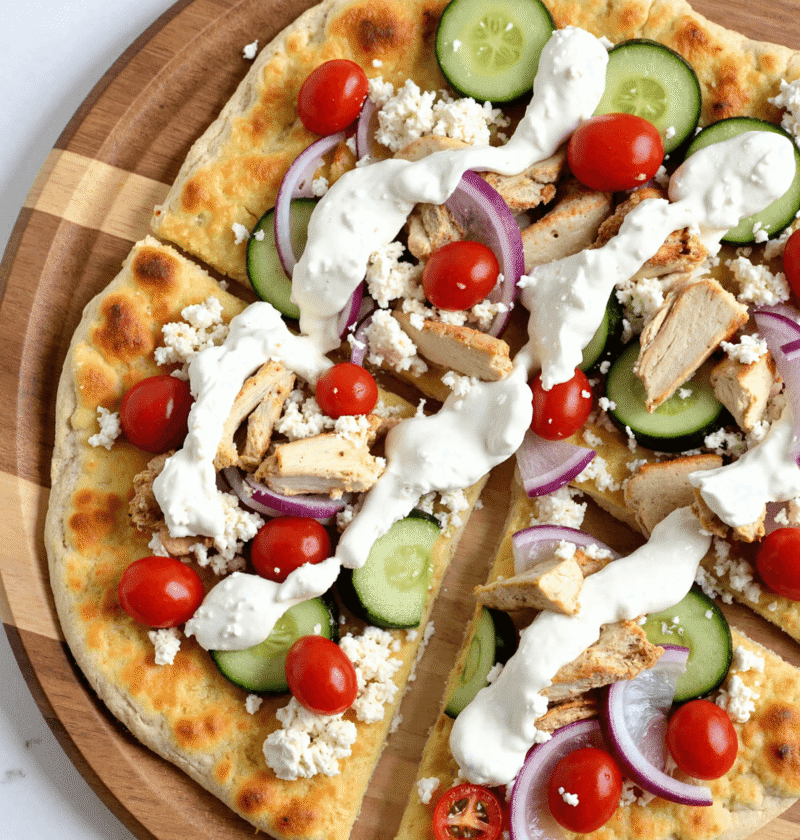 Flatbread pizza topped with grilled chicken, cherry tomatoes, cucumbers, red onion, feta, and tzatziki, sliced and served on a round wooden platter.